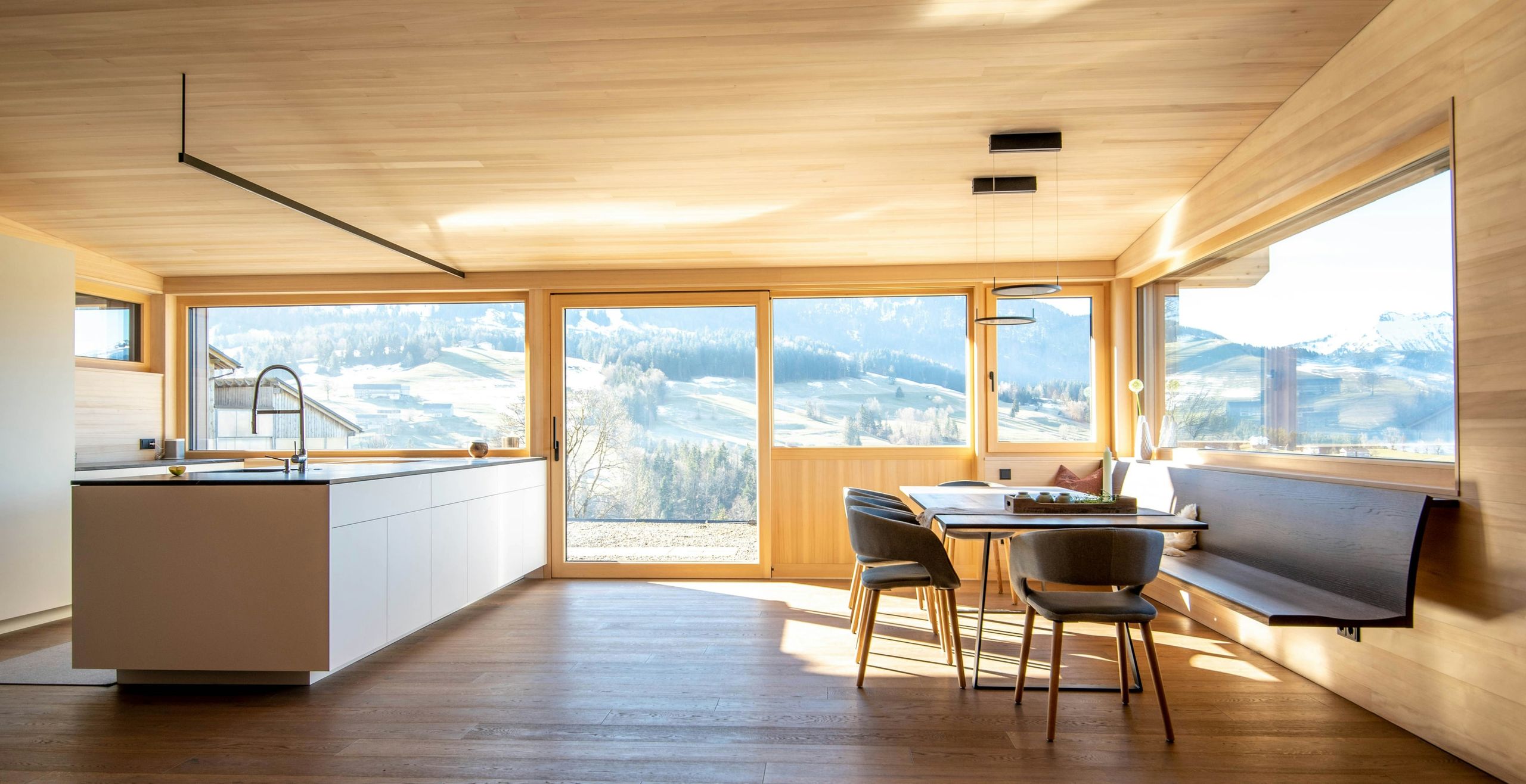 A timber-lined kitchen and dining room with wide windows onto hills
