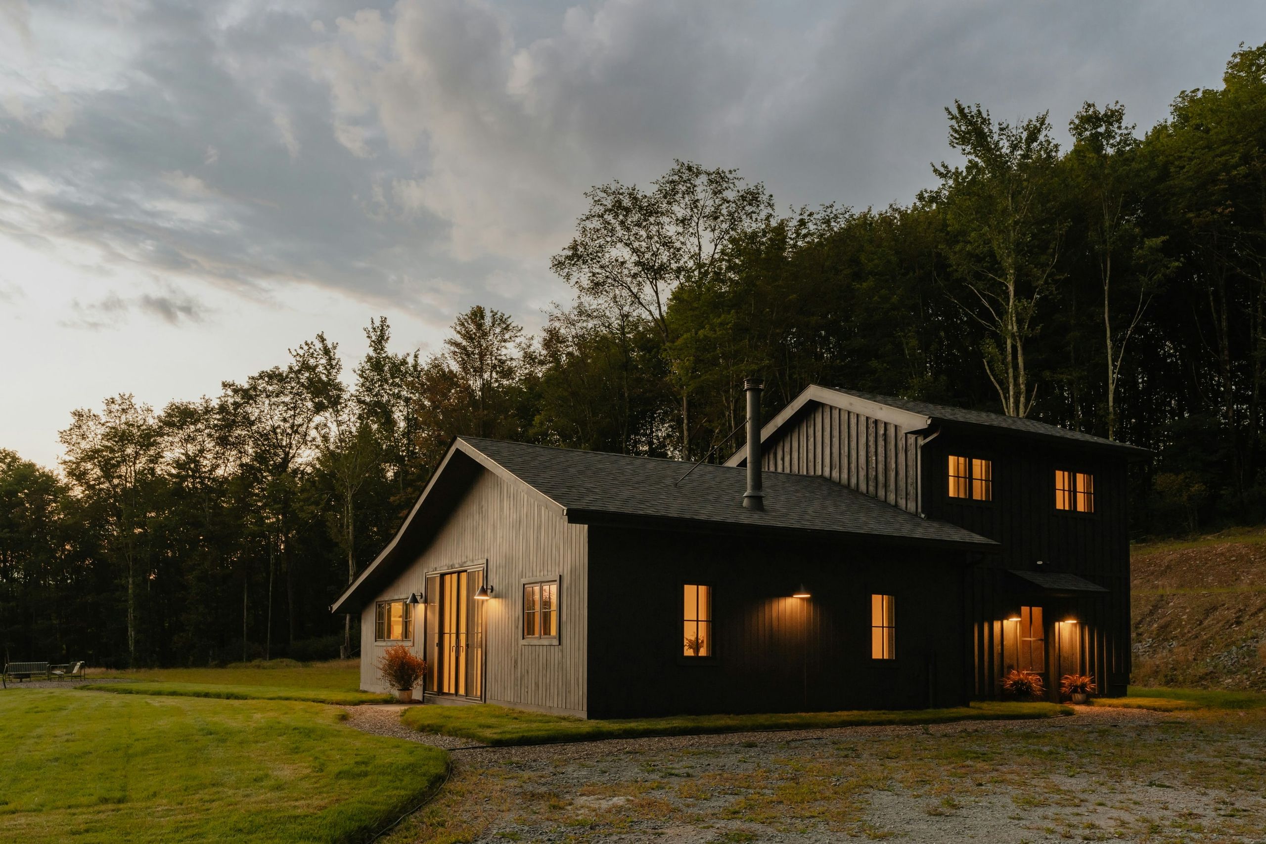 A dark timber-clad house at dusk, its windows lit warm against a stand of trees