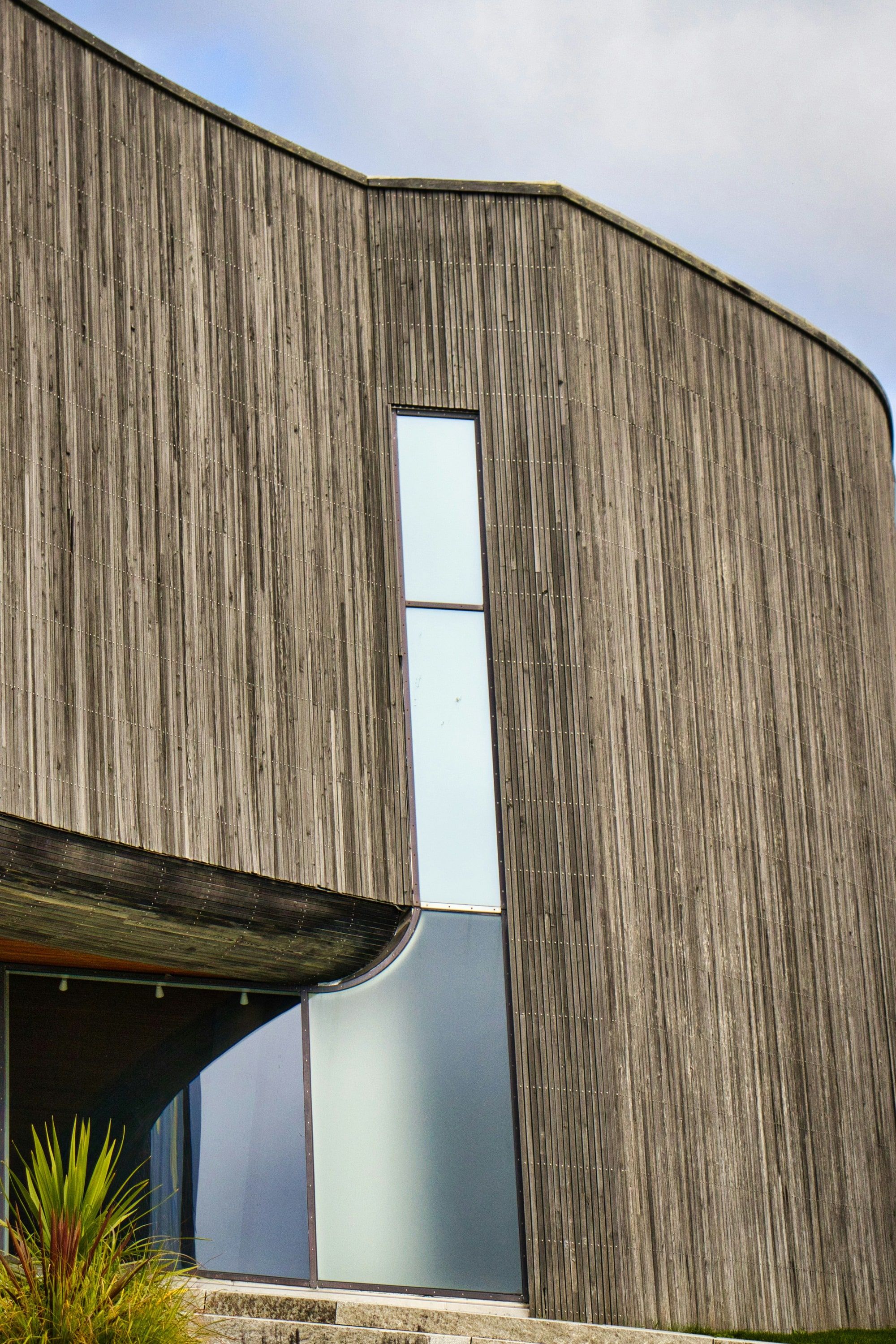 Weathered vertical timber cladding curving around a building, with a slot window