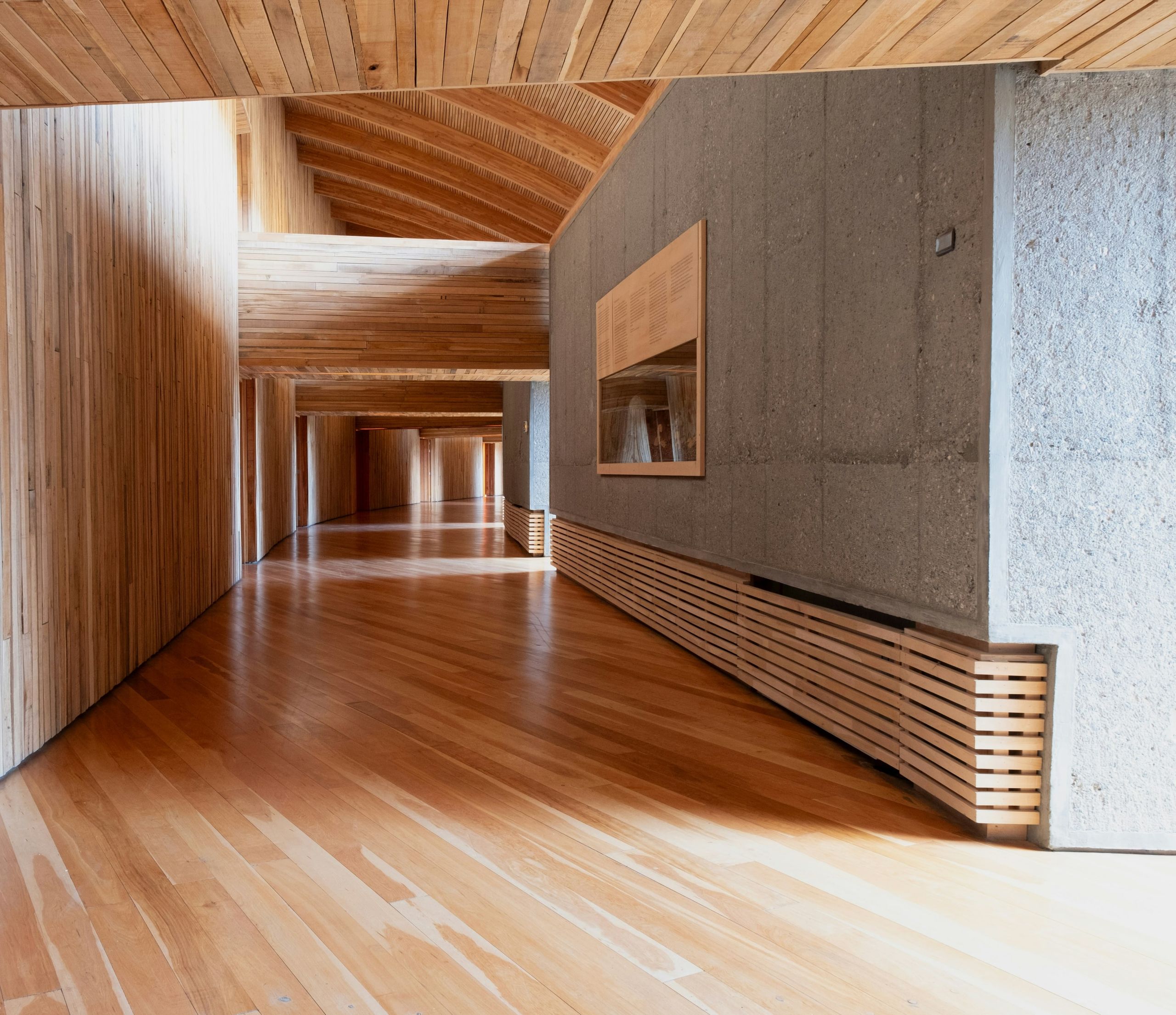 A long gallery lined in timber and board-marked concrete, light falling across the floor